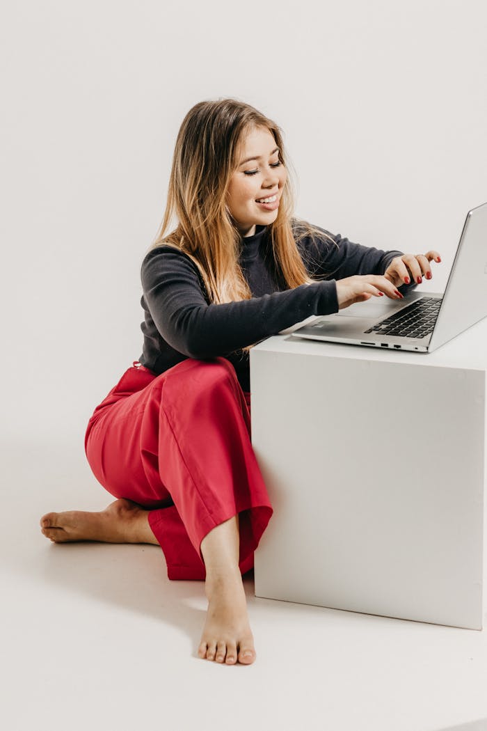 services-01 Portrait of a young woman in red trousers working on a laptop in a bright studio setting.